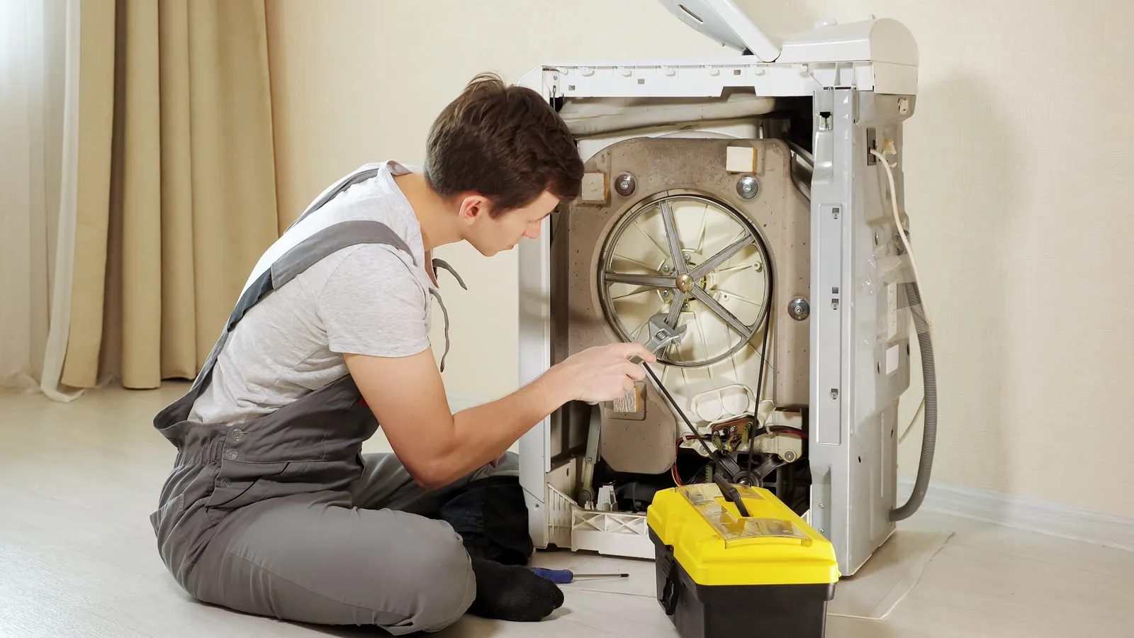 Person in grauer Latzhose an der geöffneten Rückseite einer Waschmaschine, davor ein Werkzeugkasten.